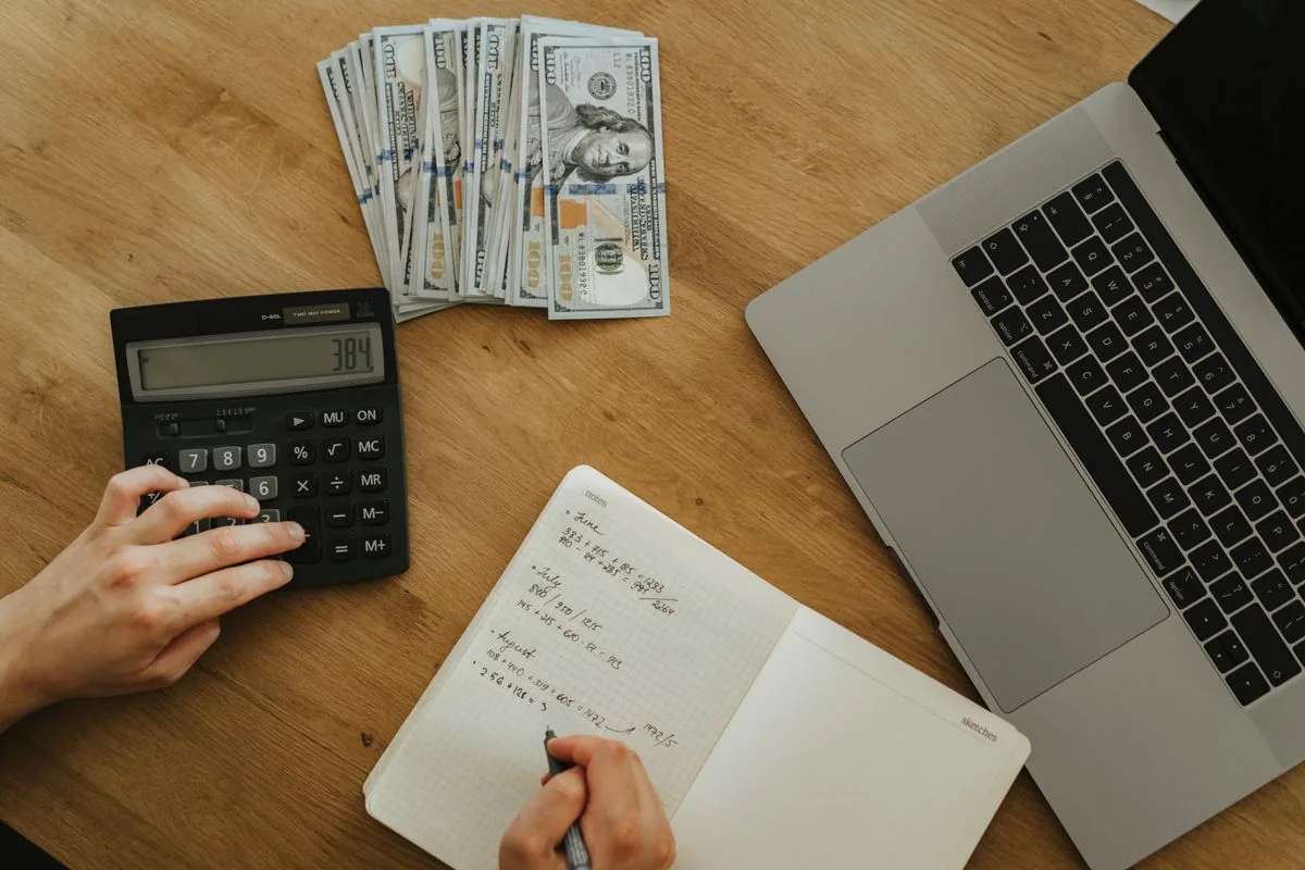 Hands using a calculator and writing in a notebook beside a laptop on a wooden table.
