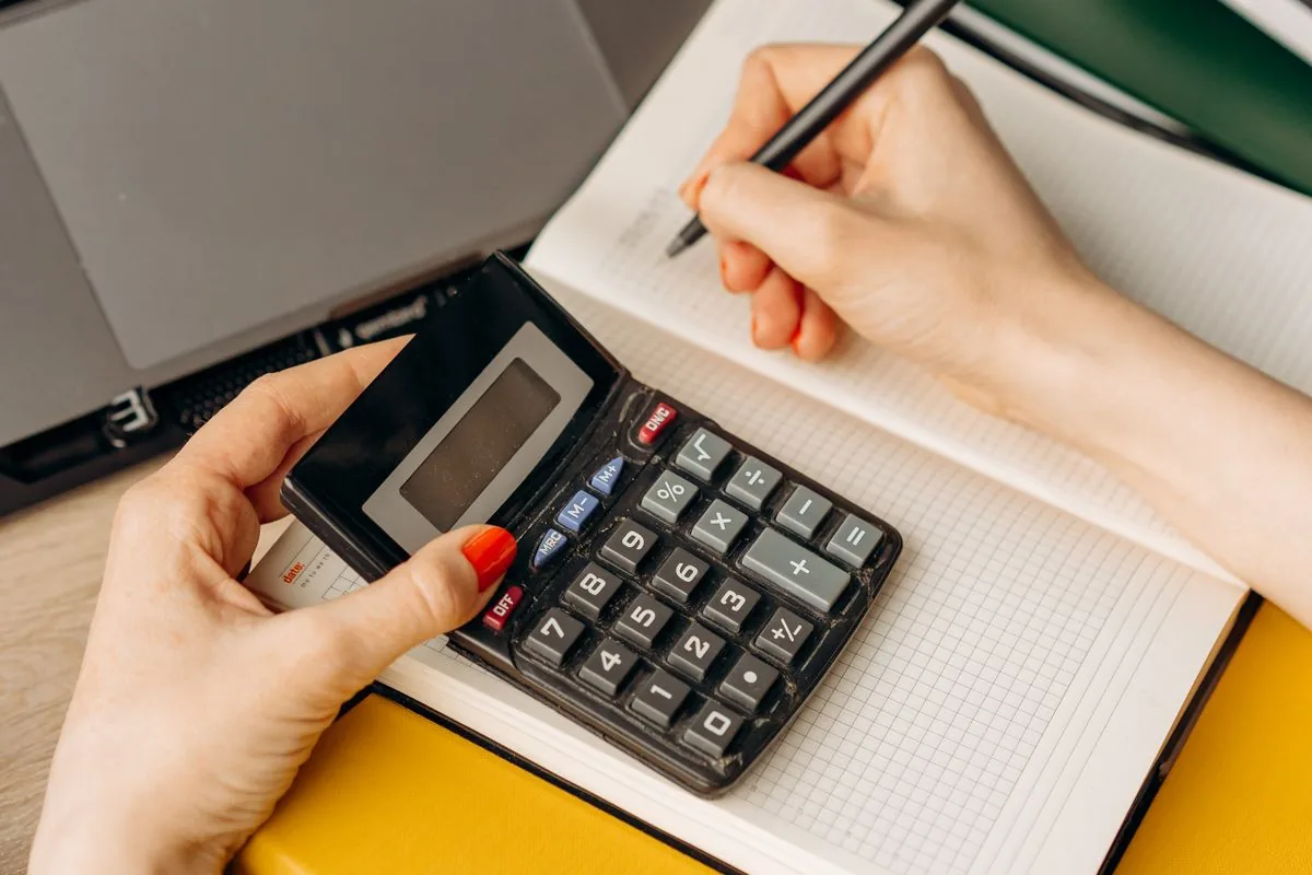 A person checking figures on a calculator and noting them in a lined notebook.