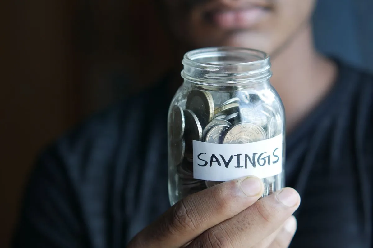 A person holding a glass jar filled with coins and labelled savings.