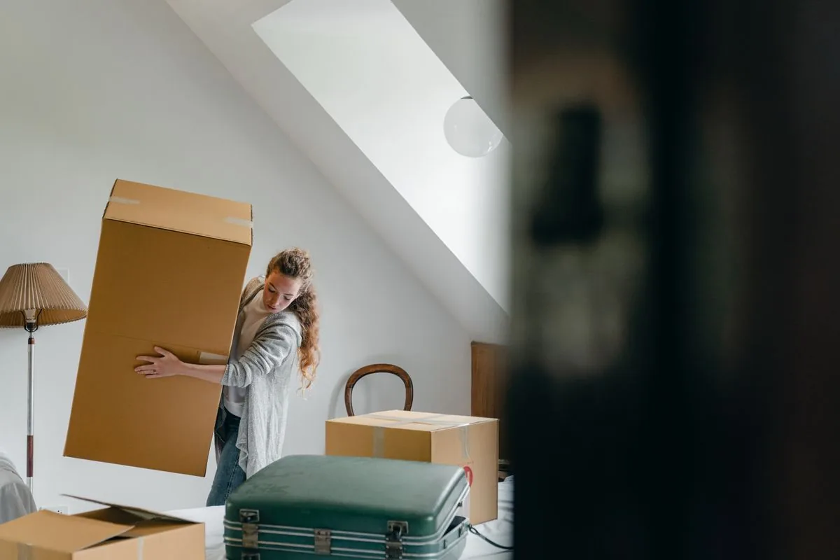 A woman carrying a cardboard moving box into a bright apartment.
