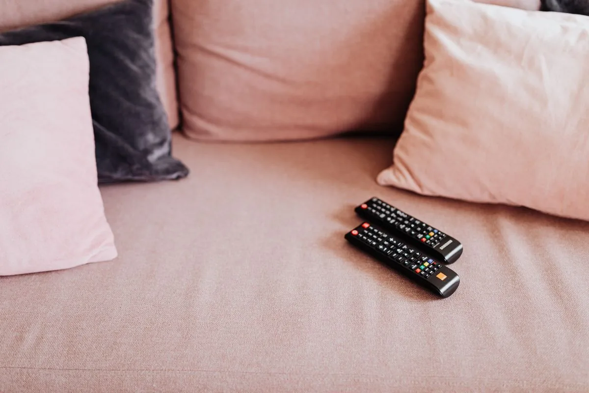 Two television remote controls resting on a soft pink sofa beside cushions.