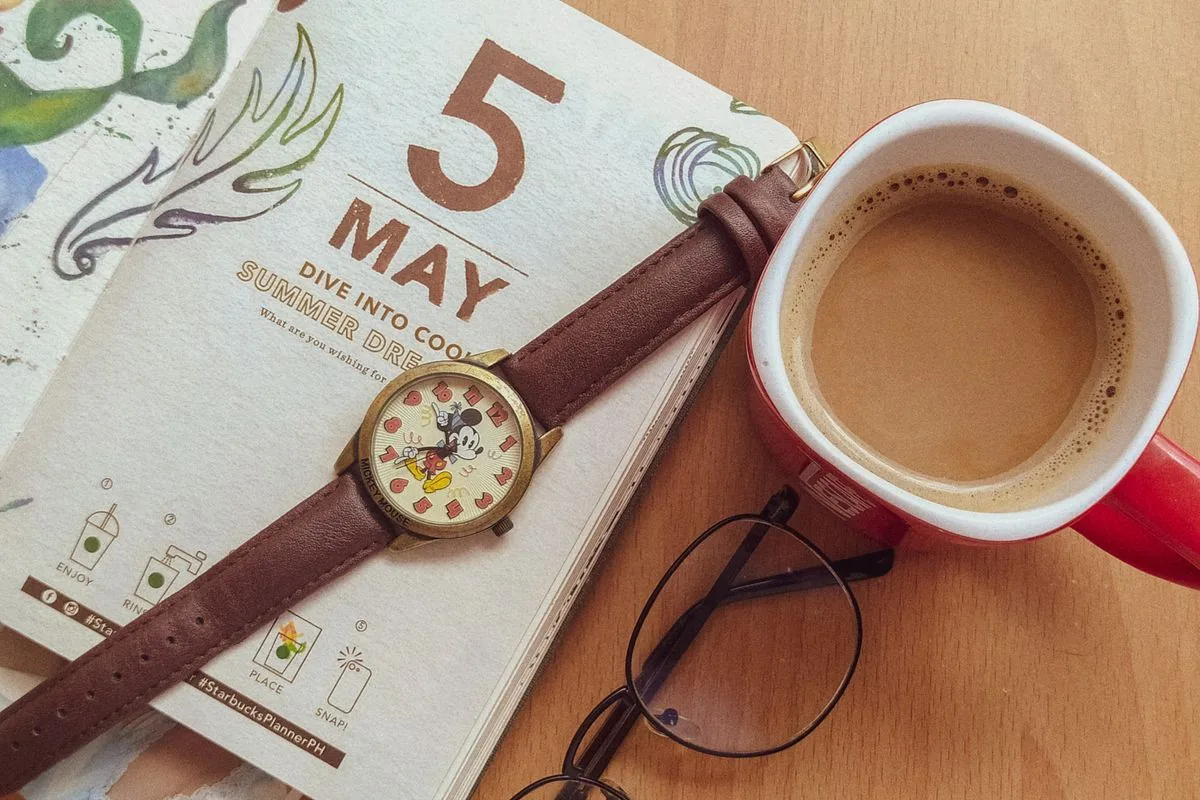 A paper planner, cup of coffee, glasses and watch arranged on a wooden desk.