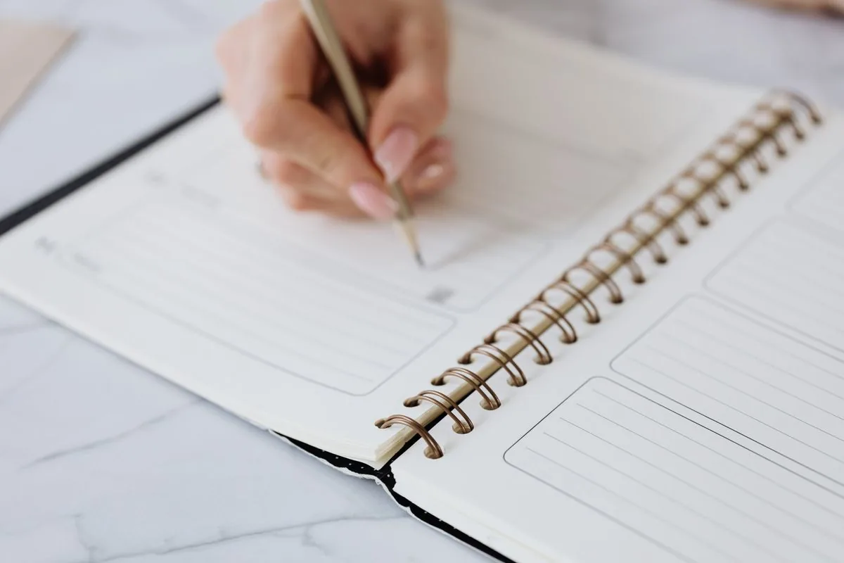 A hand writing with a pencil in an open spiral-bound notebook.