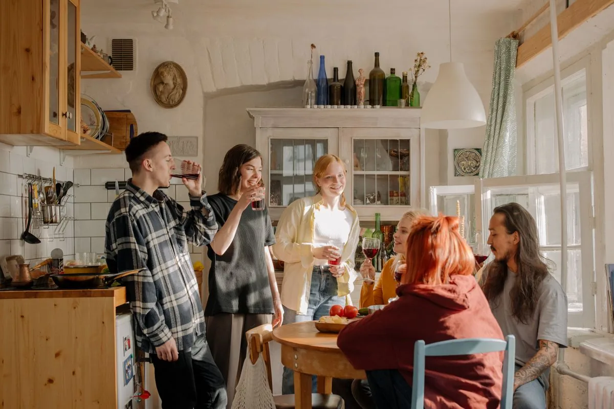 Friends talking around a small kitchen table in a sunlit apartment.