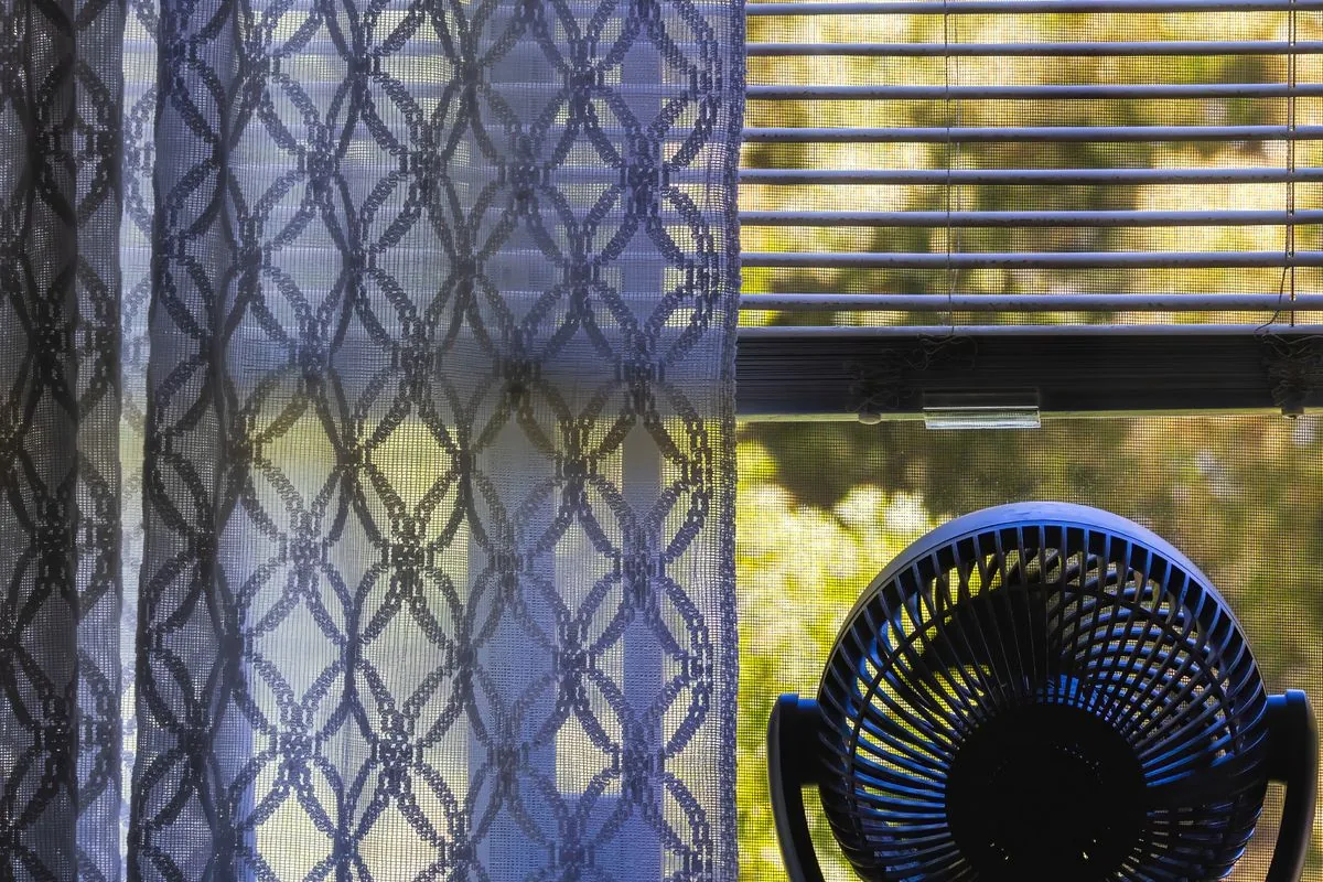 A small electric fan beside a window with blinds and patterned curtains.